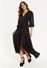 Black wrap dress made of satin with elbow-length puff sleeves, side tie detail, high slit, and flowing design. Paired with black block heels.