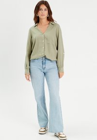 Chemise boutonnée vert olive avec col et poche unique, associée à un jean évasé bleu clair et des baskets brunes avec des accents blancs.