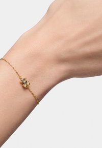 Bracciale d'oro con un elemento decorativo centrale dotato di una pietra preziosa trasparente, indossato su un polso dalla texture liscia. Design a catena delicata.
