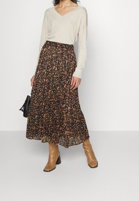 Jupe noire fluide avec imprimé floral aux tons orange et jaune, design plissé, associée à un haut beige ample à manches longues et des bottes à talons marron.