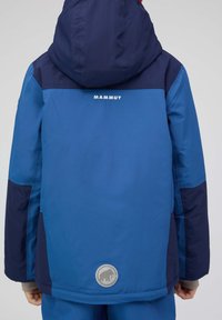 Blaue und marineblaue gefütterte Jacke mit Kapuze, verfügt über ein Logo auf dem Rücken und ein rundes Patch am Saum. Texturiertes Material, lässiges Design.