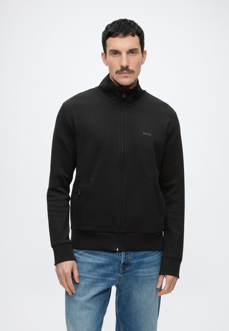 Man met kort donker haar en snor, gekleed in een zwarte jas met ritssluiting en blauwe jeans, staand tegen een effen lichte achtergrond.