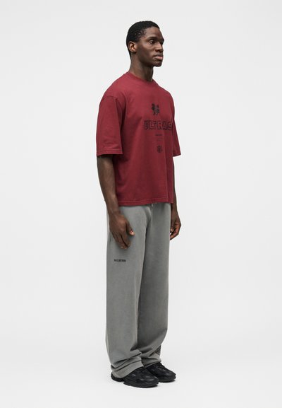 T-shirt oversized em vinho com impressão preta "ULTRA$", combinada com calças de fato de treino largas cinzentas, com um pequeno logótipo perto da bainha. Ténis pretos.