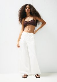 Hnedý crop top s volánovými okrajmi a malými bodkami spárovaný s širokými bielymi nohavicami a hnedými šľapkami na svetlom pozadí.