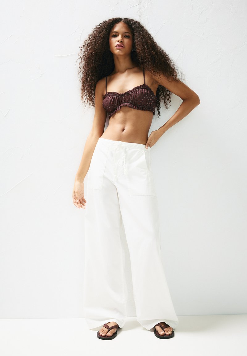 Hnedý crop top s volánovými okrajmi a malými bodkami spárovaný s širokými bielymi nohavicami a hnedými šľapkami na svetlom pozadí.