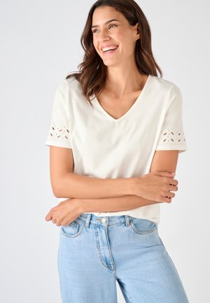 Femme souriante, portant un haut blanc à manches courtes avec des détails œillets sur les manches et un jean bleu clair taille haute, bras croisés.