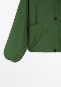 Groene jas met lange pofmouwen, voorzak en zwarte drukknoopjes op een witte achtergrond.