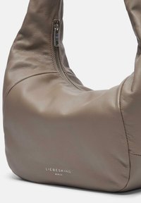 Jasnobrązowa skórzana torebka o gładkiej fakturze, zakrzywionym kształcie i zamek błyskawiczny z przodu, z subtelnym logo na dole.