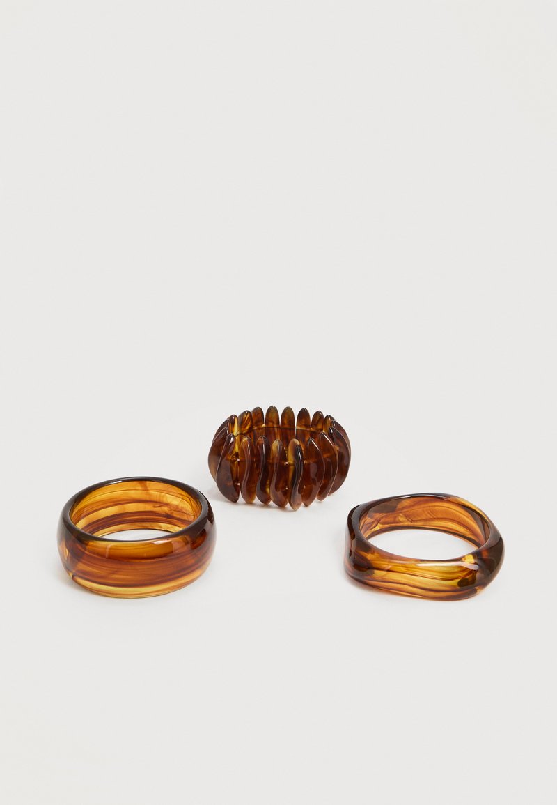 Trois anneaux en résine translucide de couleur ambre, avec des formes et textures différentes, posés sur une surface blanche.