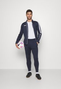 Marineblauw atletisch trainingspak met witte strepen, nauwsluitende broek en een ritsjack. Zwarte voetbalschoenen en een meerkleurige voetbal.