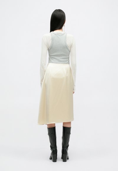 Top sin mangas gris con textura de canalé, superpuesto sobre una camiseta blanca de manga larga, combinado con una falda midi de satén color crema y botas de tacón alto negras.