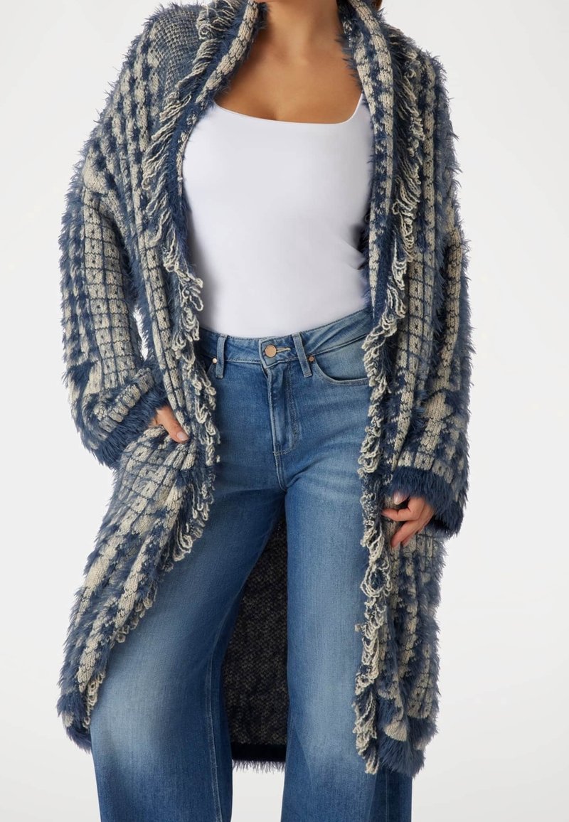 Femme portant un cardigan à franges texturé bleu et beige par-dessus un débardeur blanc et un jean bleu taille haute, les mains dans les poches du cardigan.
