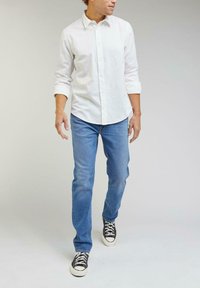 Chemise blanche à manches longues, jeans bleu clair et baskets basses noires avec des semelles blanches. Les manches de la chemise sont retroussées au niveau des poignets.
