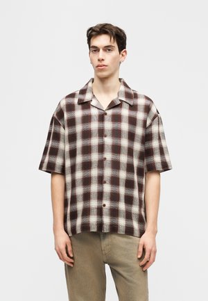 Ung mand iført en kortærmet brun og hvid ternet skjorte med beige bukser, stående mod en ensartet lys baggrund.