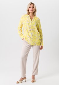 Gele bloemmotieven blouse met knoopsluiting op de kraag, lange mouwen en een gebogen zoom. Gepaard met beige rechte broek en lichte sandalen.