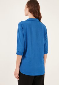 Camisa azul de manga larga con textura, cuello y mangas enrolladas, con un corte relajado y un acabado suave, combinada con pantalones negros.