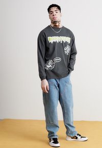 Long-sleeve black shirt with green graffiti-style text and cartoon designs, paired with light blue baggy jeans and black sneakers.
