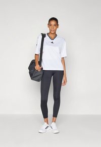 Balta sporta krekls ar melnām detaļām un īsām piedurknēm, kopā ar melnām legingiem un baltiem sporta apaviem. Komplektā ietverta pelēka plecu soma.