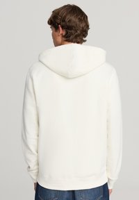 Uomo con capelli castani corti, che indossa un semplice hoodie bianco e jeans blu, rivolto di spalle su uno sfondo chiaro.