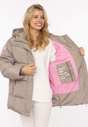 Blonde Frau lächelt, trägt eine beige Daunenjacke, geöffnet, um das pinke Futter mit dem Text "ALWAYS KEEP SMILING AND HAVE FUN." zu zeigen