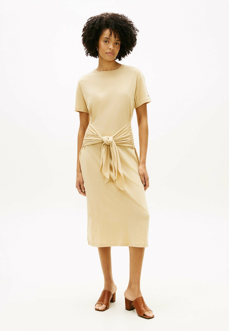 Femme aux cheveux bouclés portant une robe midi beige avec un nœud noué à la taille et des sandales à talons brunes à enfiler.