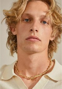 Gouden ketting met onregelmatige schakels, gedragen om de nek tegen een lichte blouse. Zachte textuur en glanzende afwerking.
