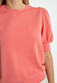 Sudadera de color coral con mangas cortas abullonadas, cuello redondo y textura suave. Presenta un corte relajado y un brillo sutil.