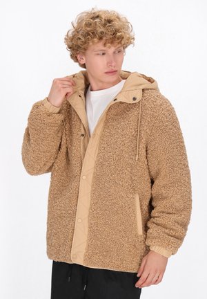 Tânăr ajustând gulerul unei jachete din fleece sherpa bej cu glugă matlasată și butoane prin presare, îmbrăcând o cămașă albă și pantaloni negri.