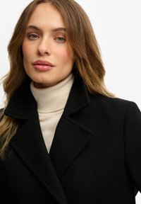 Zwarte wollen jas met een diepe kraag, gedragen over een crèmekleurige turtleneck. Gladde textuur, slim fit ontwerp en strakke lijnen.