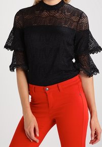 Sort blondebluse med korte flæser og høj hals, kombineret med røde tætsiddende jeans med forlomme og knapdetalje.