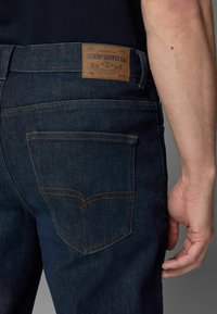 Tumedatest sinisest denimsest teksad, millel on pruun nahk silt õmmeldud vööle, tagaküljel on kotid, millel on kontrastne õmblus.