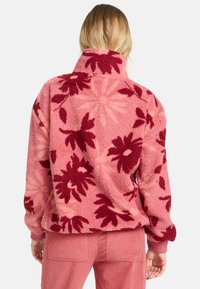 Fleece-Pullover in Pink mit maroonfarbenem Blumenmuster, ausgestattet mit einem hohen Kragen und gerippten Bündchen. Weicher Stoff und lockere Passform.