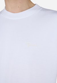 Hvit bomulls T-skjorte med ribbet rund hals, med en subtil, lys gull brodert logo på venstre brystområde.
