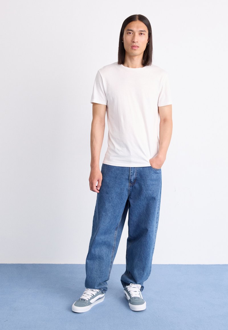 Hombre con cabello oscuro y largo, vistiendo una camiseta blanca, jeans azules sueltos y zapatillas grises, de pie con una mano en el bolsillo sobre un suelo azul.