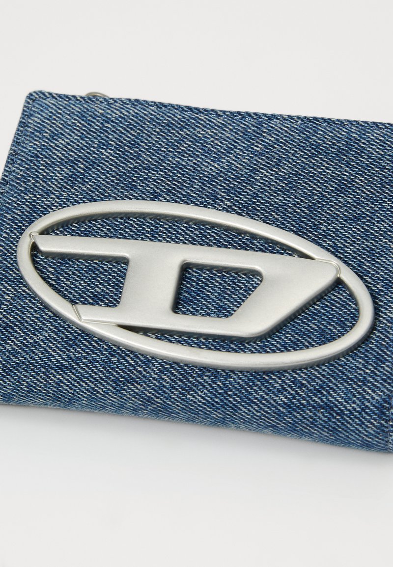 Borsa a mano in denim con un grande logo metallico argentato a forma ovale. Il tessuto presenta una finitura in denim blu texturizzato. Chiusura con zip nella parte superiore.