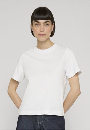 Donna con capelli corti e scuri che indossa una maglietta bianca semplice a maniche corte e jeans scuri su uno sfondo neutro.