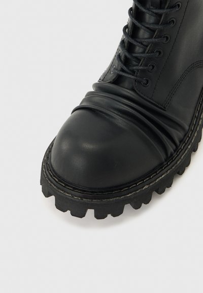 Μαύρο μποτάκι από δέρμα με κορδόνια, χοντρή σόλα με ανάγλυφα πέλματα και ραμμένες λεπτομέρειες, εμφανίζεται από κοντινή γωνία μπροστά.