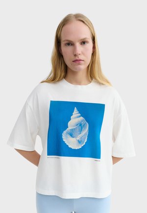 Młoda kobieta z długimi blond włosami nosi białą, oversize'ową koszulkę z dużym niebieskim kwadratem i białym nadrukiem muszli.