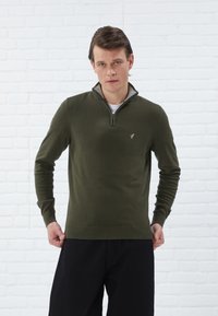 Pull en tricot vert olive avec un col montant et une demi-fermeture éclair, présentant un logo discret sur la poitrine ainsi que des poignets et un ourlet côtelés.