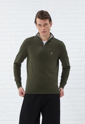 Pull en tricot vert olive avec un col montant et une demi-fermeture éclair, présentant un logo discret sur la poitrine ainsi que des poignets et un ourlet côtelés.