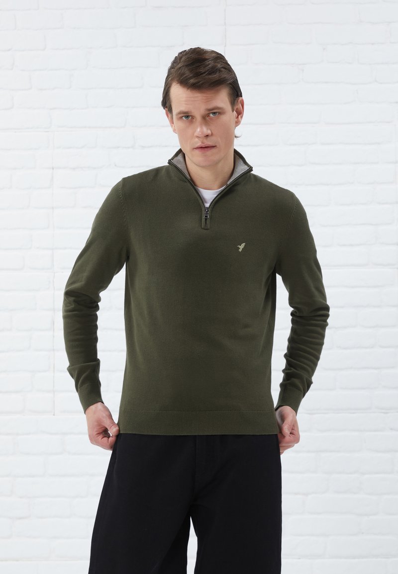 Pull en tricot vert olive avec un col montant et une demi-fermeture éclair, présentant un logo discret sur la poitrine ainsi que des poignets et un ourlet côtelés.