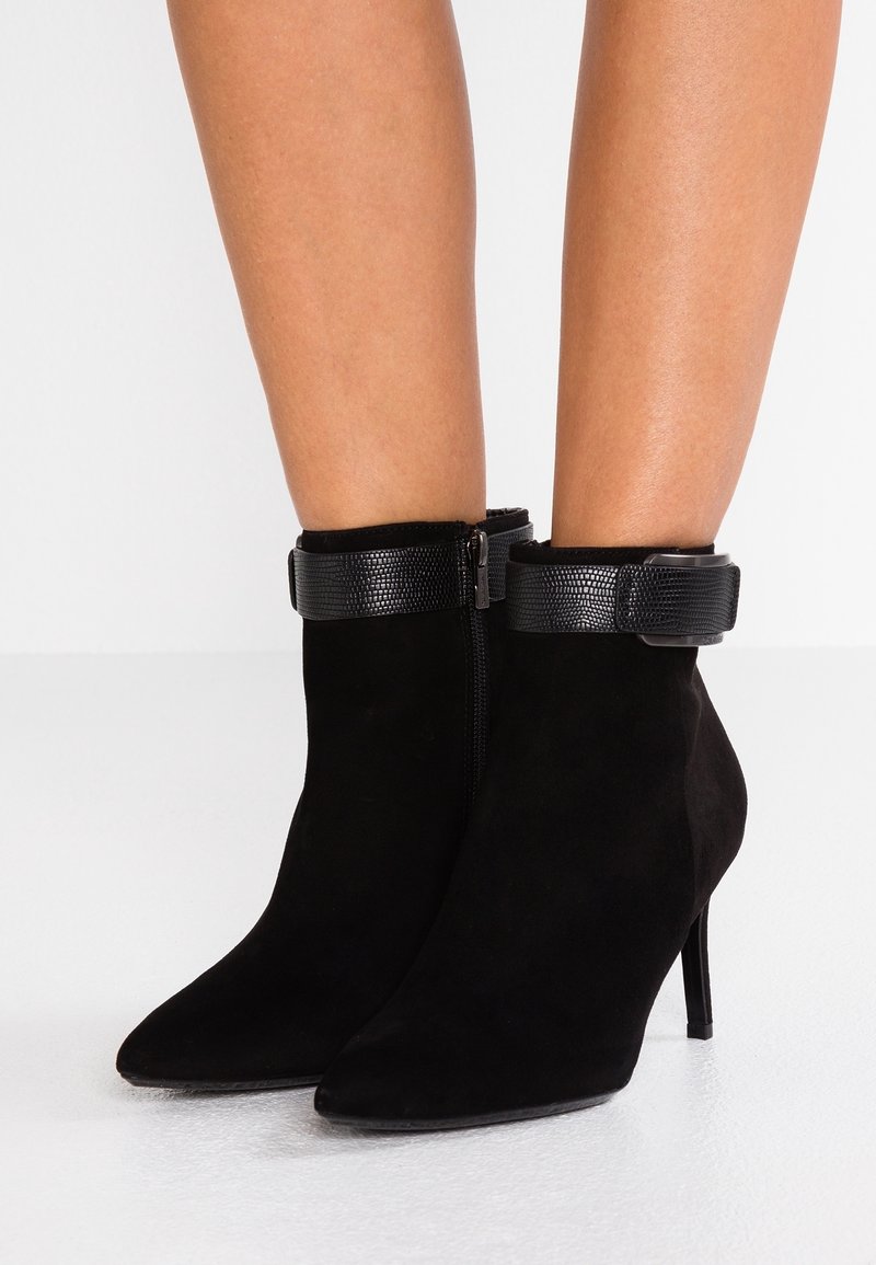 Bottines en daim noir avec un bout pointu et un talon stiletto haut. Dotées d'une sangle noire texturée et d'une fermeture éclair sur le côté.