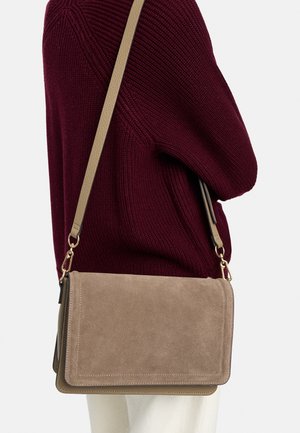 Beżowa torebka na ramię z zamszu z złotymi okuciami, noszona przez osobę w bordowym swetrze z dzianiny i białych spodniach, widziana z boku.