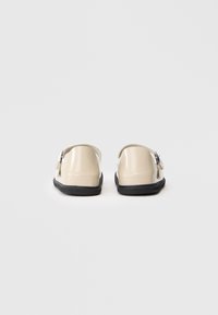 Scarpe beige slip-on con suole nere e cinturini laterali a fibbia, viste da dietro su uno sfondo bianco uniforme.