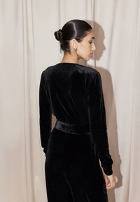 Abito lungo in velluto nero con maniche lunghe, dalla silhouette aderente, dotato di una cintura in vita e un leggero lucido texturale, ambientato su uno sfondo beige.