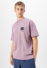 Noormees, kes kannab lavendlit värvi ülemõõdulist T-särki, millel on rinna peal moonutatud must ja oranž logo, on sinistes pükstes ja seisab valge taustal.