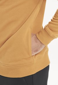 Sennepsgul sweatshirt lavet af blødt materiale, med en sidelomme, ribbede manchetter og en buet kant. Sorte bukser er delvist synlige.