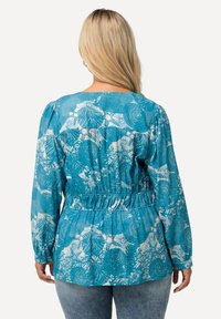 Donna con lunghi capelli biondi che indossa una blusa a maniche lunghe bianca e turchese con vita arricciata, abbinata a jeans blu sbiaditi.