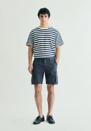 Joven de pie de frente, vistiendo una camiseta de rayas blancas y azul marino, pantalones cortos cargo oscuros y zapatos negros con cordones sobre un fondo liso.