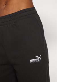 Melni sporta bikses ar elastīgu jostu un baltu Puma logo izšuvumu kreisajā augšstilbā. Mīksta auduma faktūra, ikdienišķs dizains.